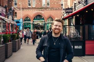 Craig Moran smiling on a busy street with brick buildings and shops in the background. People walking around, string lights above create a lively vibe. In Stitches Comedy Club, Dublin, Ireland.