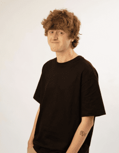 Rob Nother with curly hair and a slight smile stands against a plain background. He's wearing a black T-shirt, beige pants, and has a cartoon fish tattoo on his forearm.